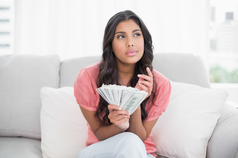 Thoughtful brunette sitting on couch holding money in bright living room